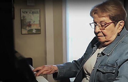 Older woman wearing glasses and blue jacket playing the piano.