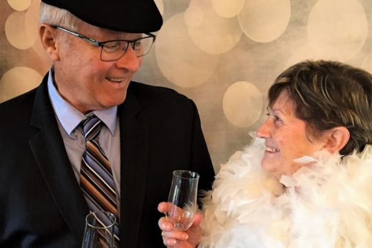 Senior couple, man wearing black suit and hat, woman in white feather boa, looking at each other and holding champagne.