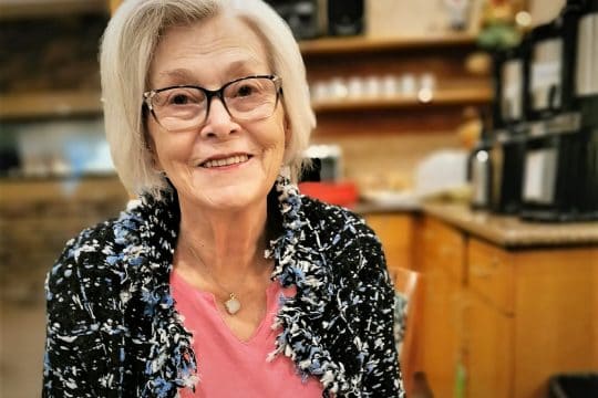 Woman with grey hair and glasses wearing a pink t-shirt and blue flowered blouse.