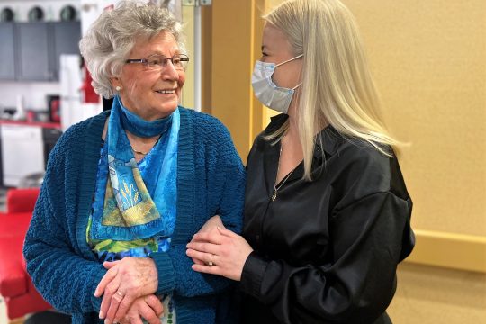 Older lady with short hair and glasses, wearing a blue patterned silk scarf looking at a young woman with long blond hair; elbows linked and both smiling.
