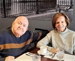 Senior man in brown and blue sweater with his arm around an older woman with dyed red hair, smiling.