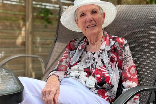 Senior woman in white had and pants, flower patterned shirt, sitting on a patio with legs crossed