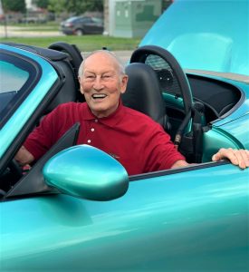 Senior man with white hair wearing a red golf shirt sitting in a blue sports care, laughing
