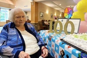 Woman with white hair sitting next to a birthday cake and a gold 100 centrepiece